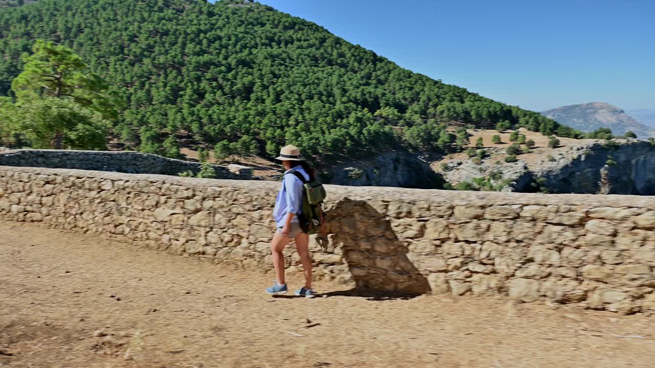 一个女人背着背包沿着风景秀丽的山路徒步旅行，在晴朗的蓝天下享受大自然和宁静的森林景观视频素材