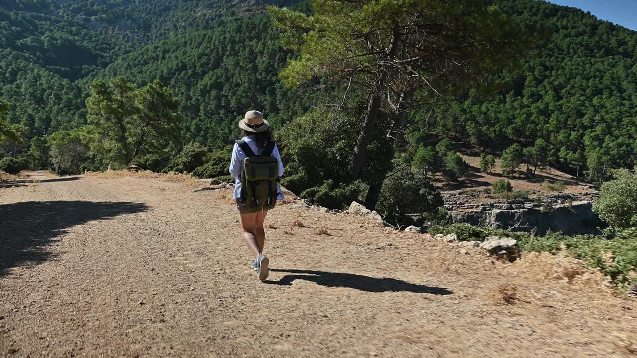 一名女子背着背包，戴着帽子，在碧绿的山间徒步旅行，在晴朗的天空下享受着大自然的宁静视频素材