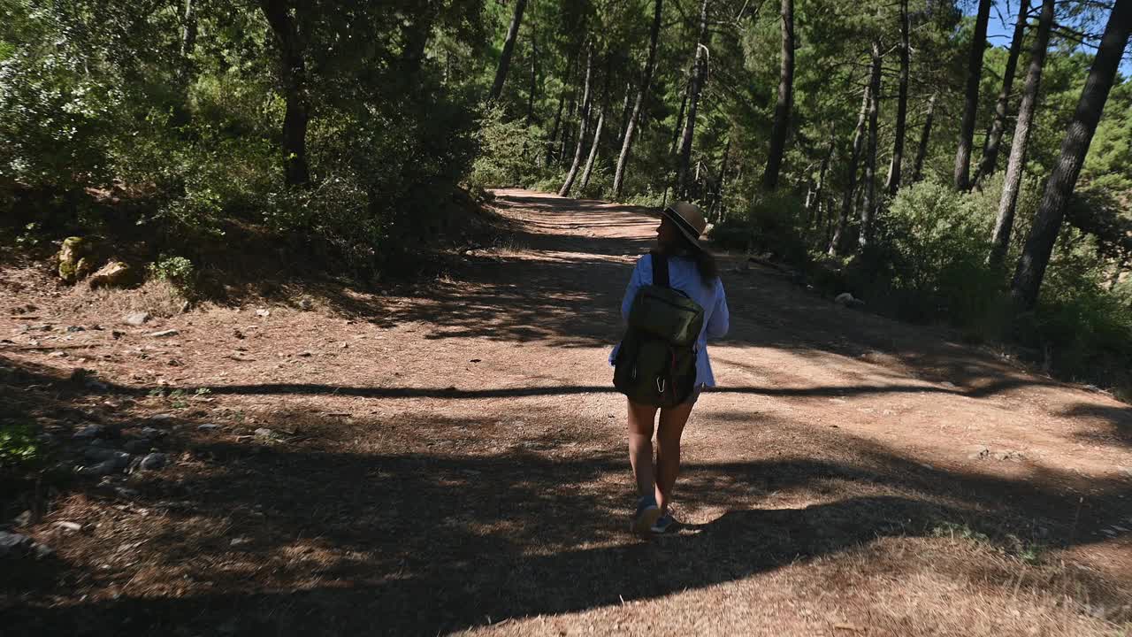 在阳光明媚的日子里，一名女子背着背包，戴着帽子，在森林小路上徒步旅行，探索大自然，享受树林的宁静视频素材