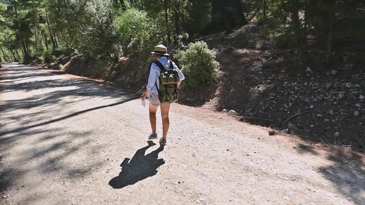 一名女子戴着帽子，穿着短裤，沿着阳光普照的森林小径徒步旅行，在夏日里背着背包探索大自然视频素材