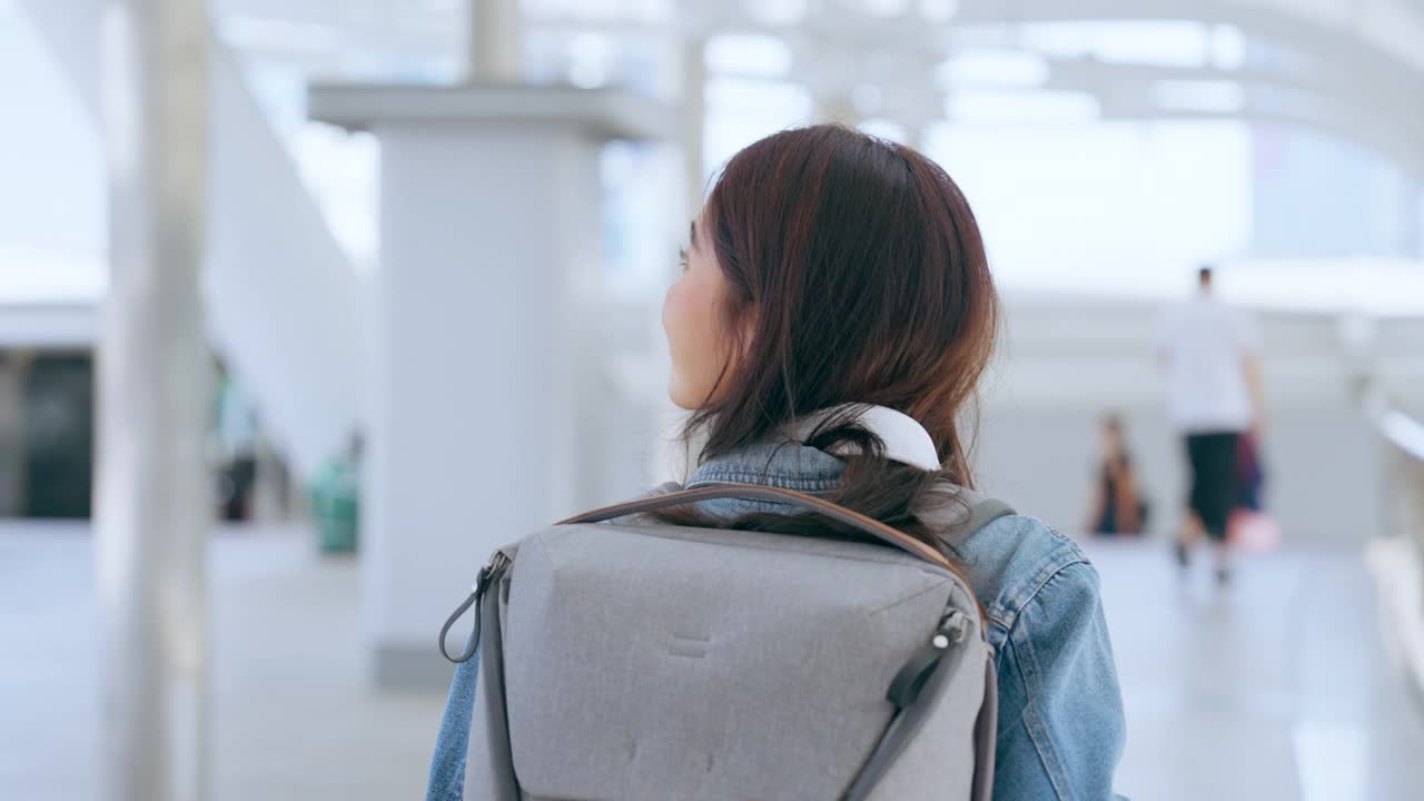 年轻的亚洲女子穿着牛仔夹克，脖子上戴着耳机，背着背包走在城市里。青春期的女性开心，笑脸盈盈。放松和享受城市生活的年轻女士视频下载