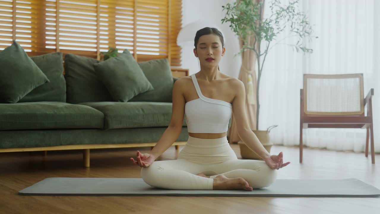 快乐的年轻亚洲女性在家里练习瑜伽和冥想，坐在客厅的地板上，以莲花的姿势放松。正念冥想，健康，呼吸练习视频下载