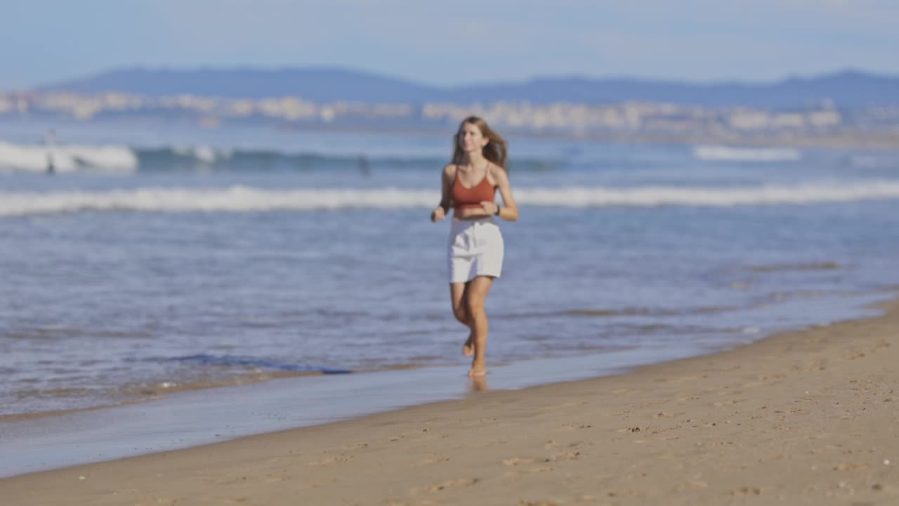 精力充沛的年轻女子沿着阳光明媚的海滩海岸线冲刺视频下载