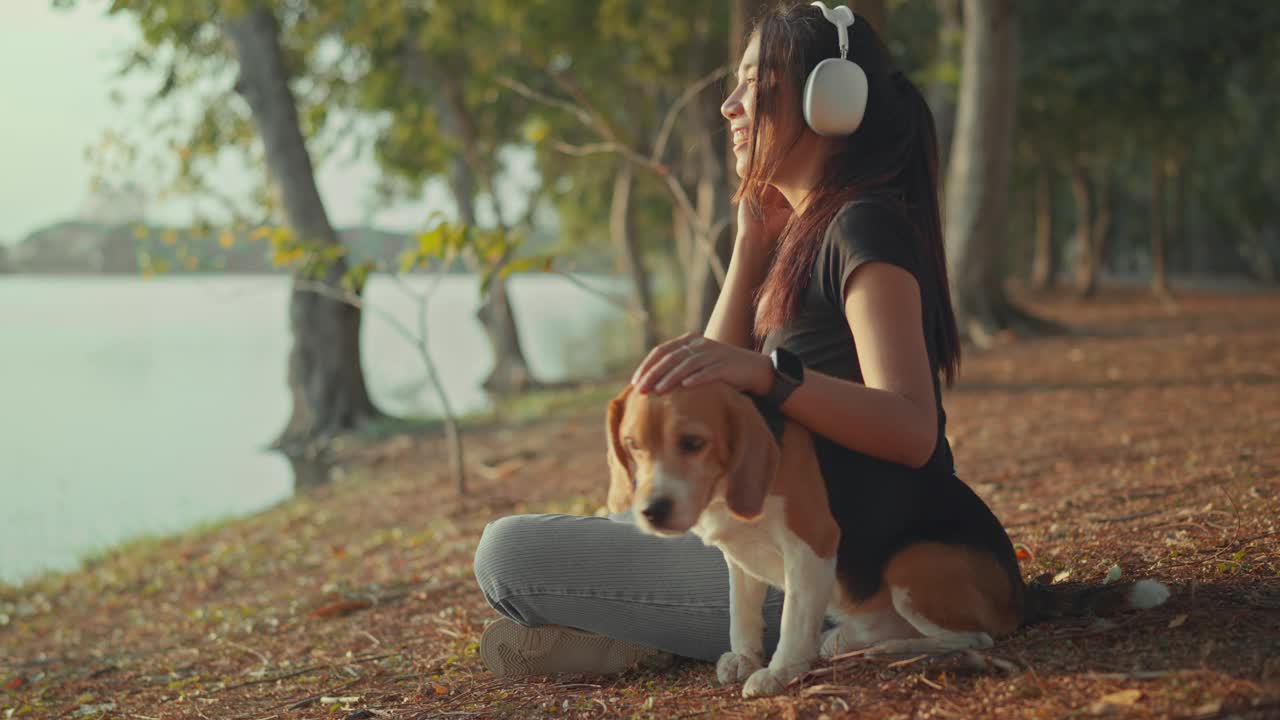 快乐的亚洲女人戴着耳机听着音乐，和小猎犬宠物一起放松，在户外夕阳下俯瞰美丽的湖泊，女孩们周末和宠物一起在大自然中度过视频下载