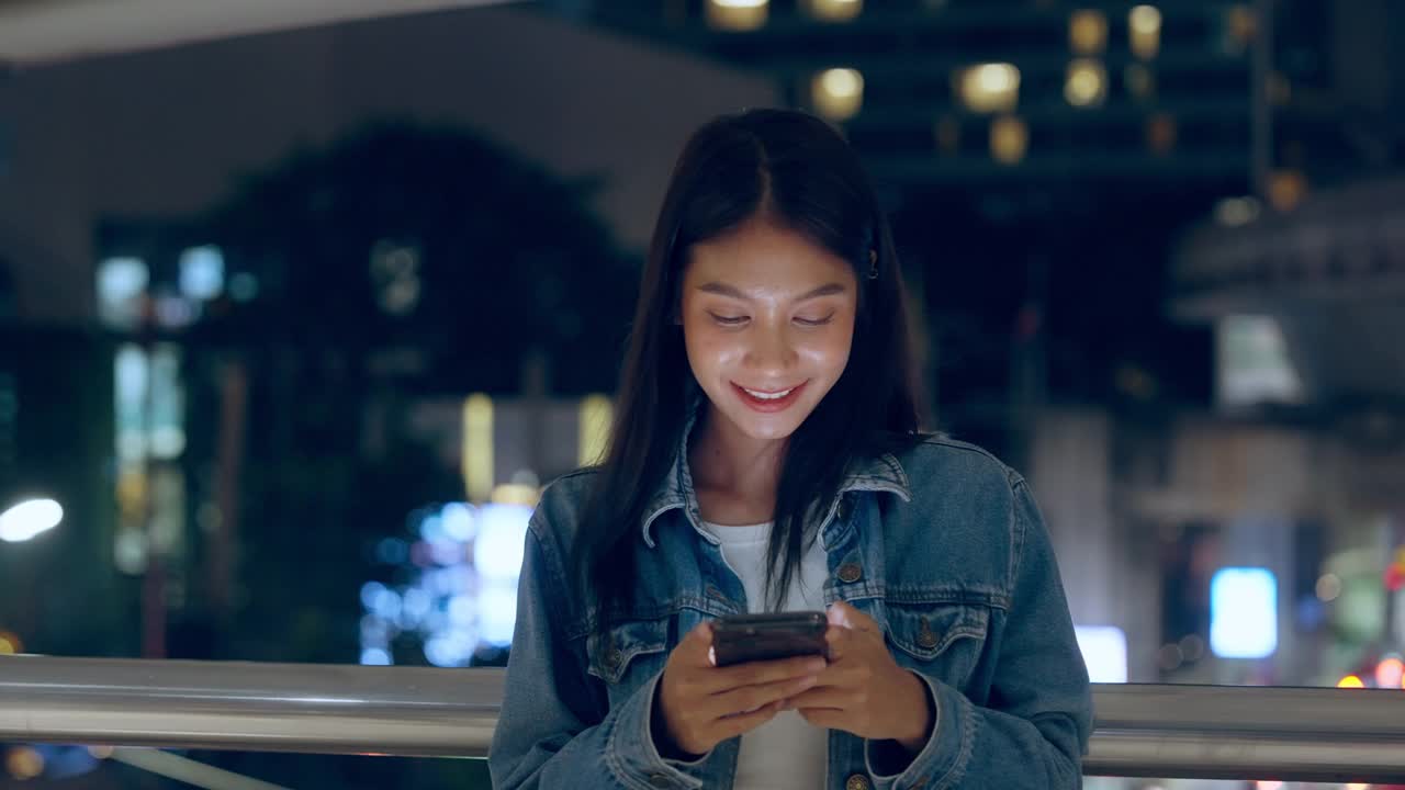 美丽的年轻亚洲女子站在城市街道在晚上使用手机。一个快乐的女人站在那里用手机打字视频下载