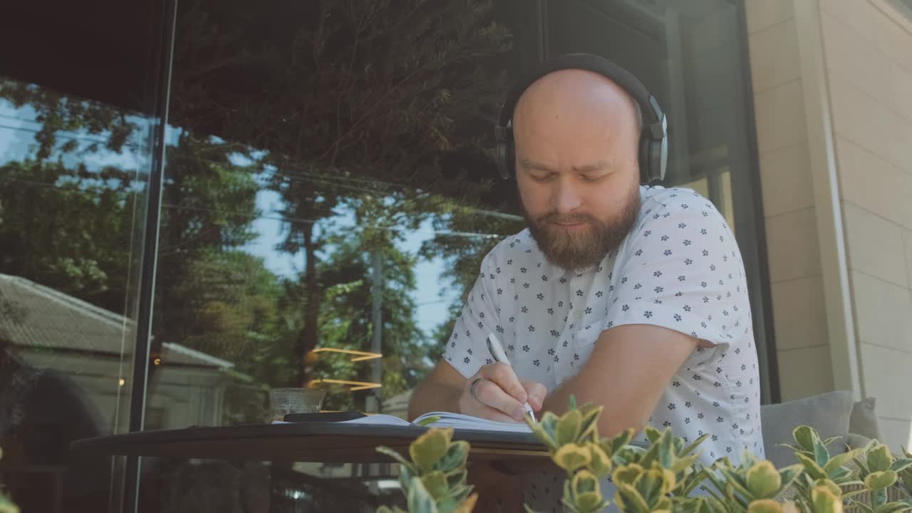 一名男子戴着耳机，坐在绿树成荫的玻璃墙旁，专注地在户外写作视频下载