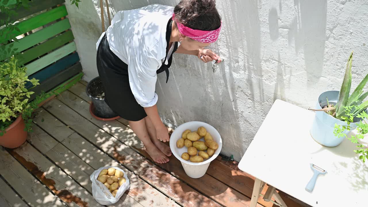 一名妇女在阳光明媚的露台上准备新鲜土豆，周围是盆栽植物和质朴的装饰，享受着一种家庭式的户外烹饪体验视频素材