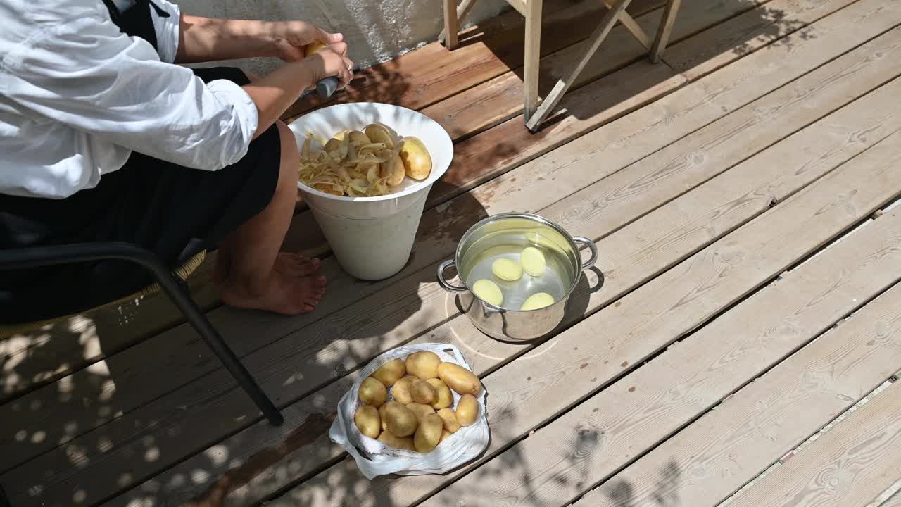 一名妇女在阳光明媚的木甲板上削土豆皮，准备做饭用的食材，唤起了一种质朴的简约感和烹饪传统。视频素材