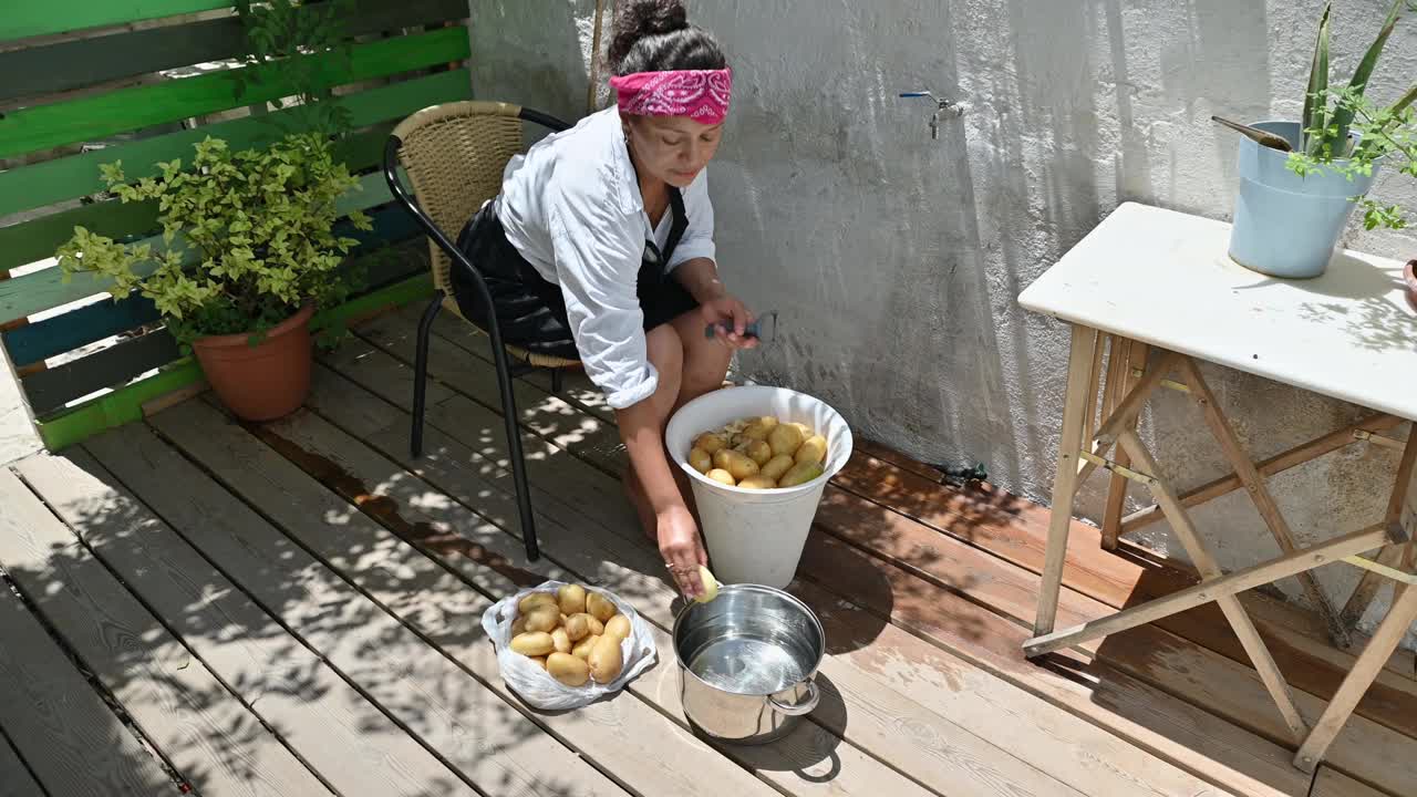 在一个阳光明媚的日子里，一名妇女在户外削土豆皮，在乡村的环境中准备一顿传统的晚餐，展示了家庭烹饪和简单视频素材