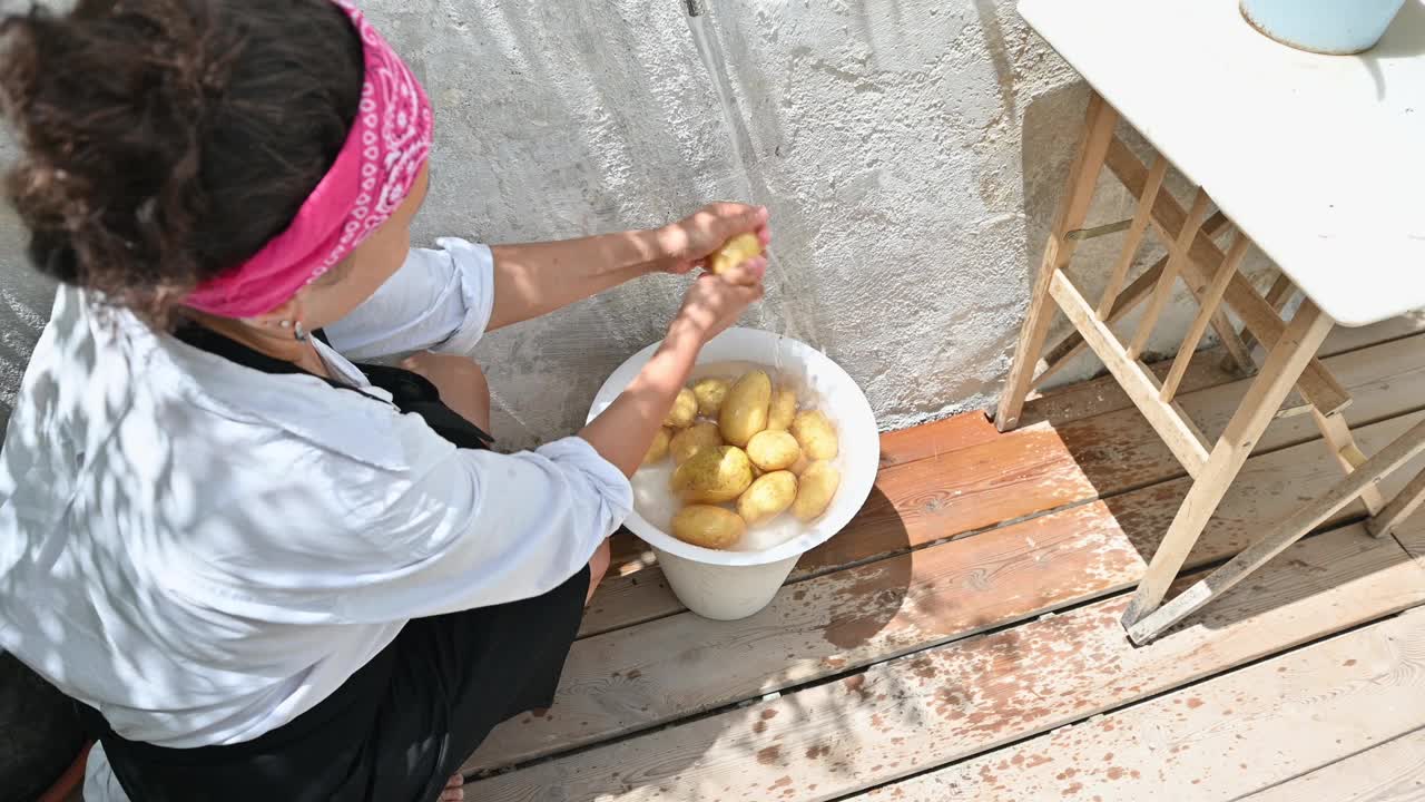 一名戴着粉色头巾的妇女在户外的木制露台上用白色的桶清洗新鲜的土豆，准备进行自制烹饪视频素材