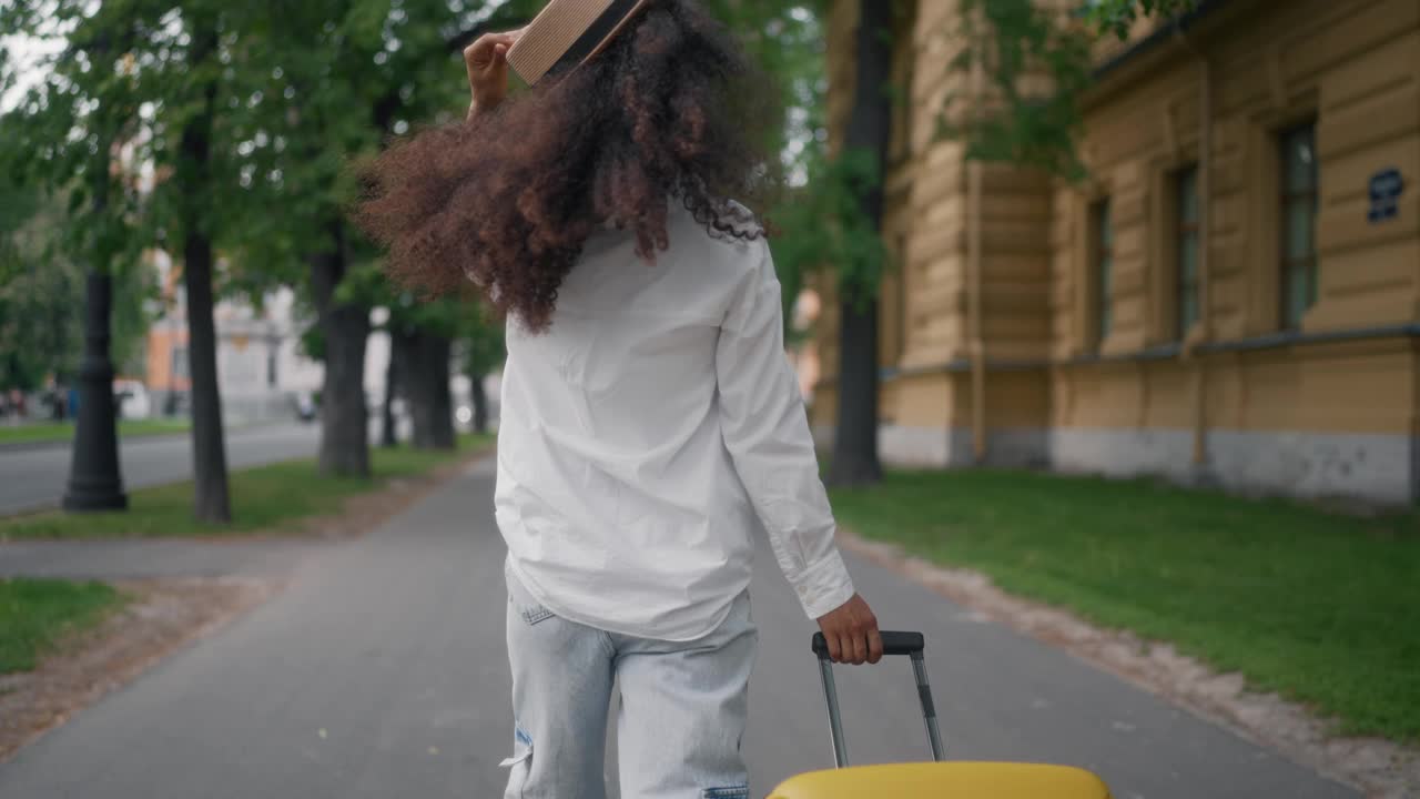 暑假周末旅行，女游客在街上滚动旅行箱。戴草帽的非洲裔美国女士独自行走后视图，观光游览游，旅游旅游概念视频素材