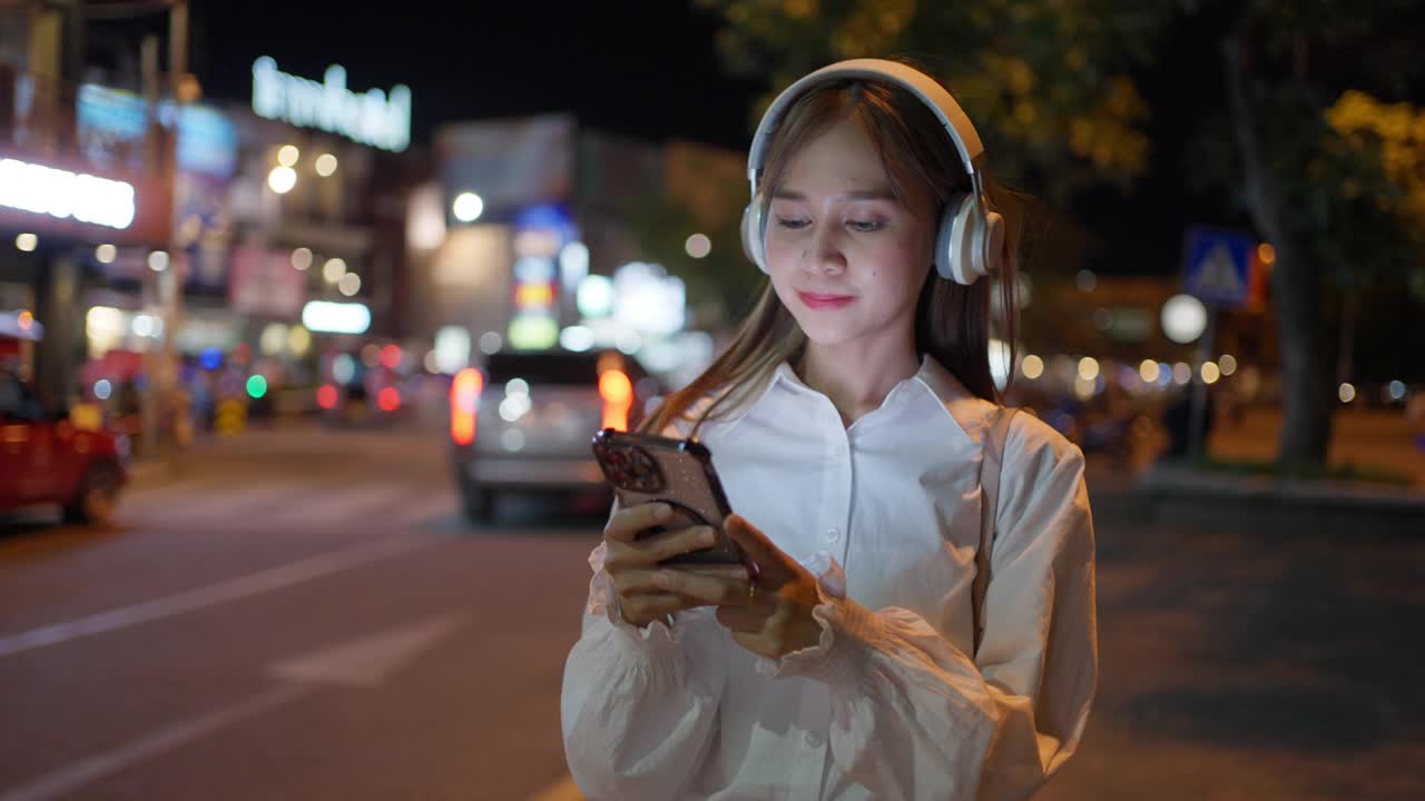 快乐的年轻女子晚上走在城市里，戴着耳机听着音乐，手里拿着手机播放着歌曲视频素材