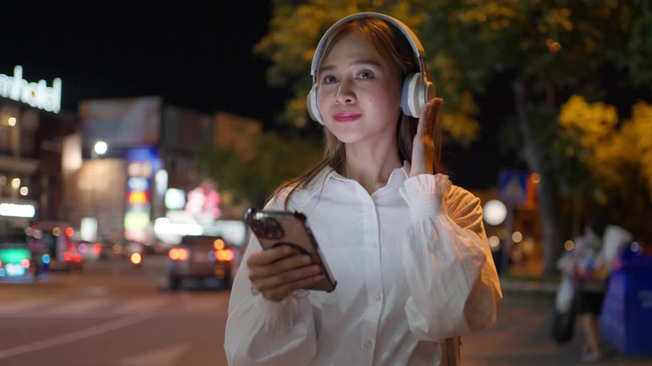 快乐的年轻女子晚上走在城市里，戴着耳机听着音乐，手里拿着手机播放着歌曲视频素材