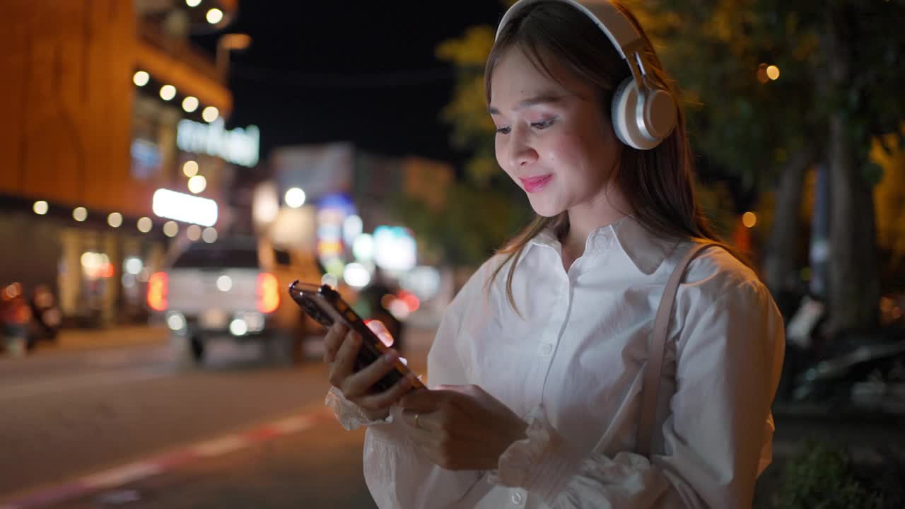 快乐的年轻女子晚上走在城市里，戴着耳机听着音乐，手里拿着手机播放着歌曲视频素材