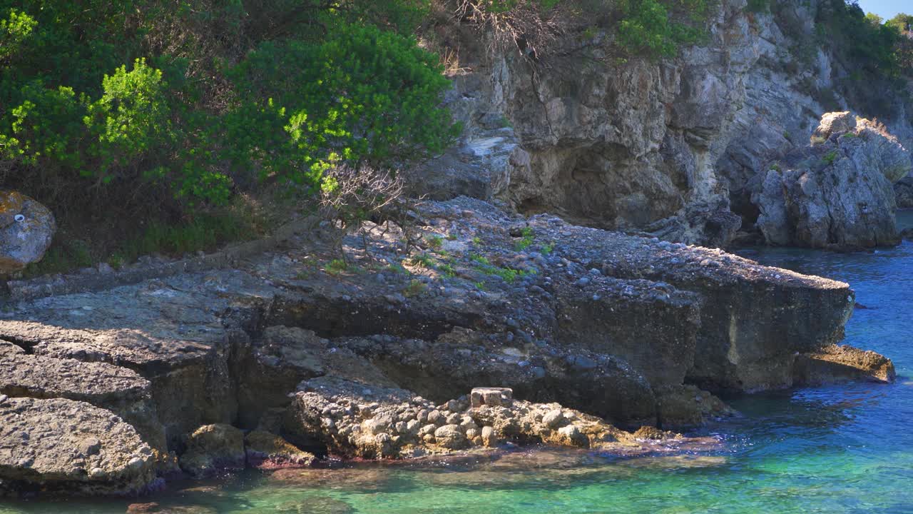 相当平静清澈的绿色大海在岩石海滩岸边，阳光照射到小波浪，典型的风景在利亚帕德斯，希腊的许多海湾之一视频下载