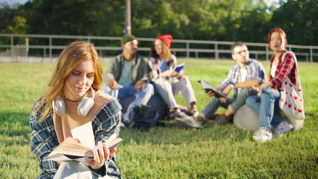 在阳光明媚的大学公园里，一名红发女学生穿着衬衫和牛仔裤，戴着耳机坐在草地上，周围是拿着学习资料的各种各样的同学视频下载