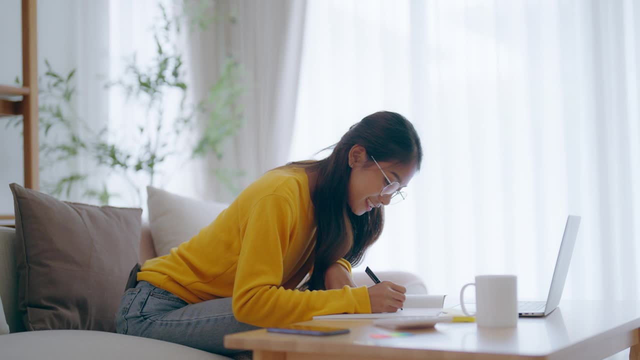 年轻的亚洲女性戴着眼镜使用笔记本电脑，在客厅工作，咖啡杯放在桌子上。舒适的办公场所，远程办公视频下载