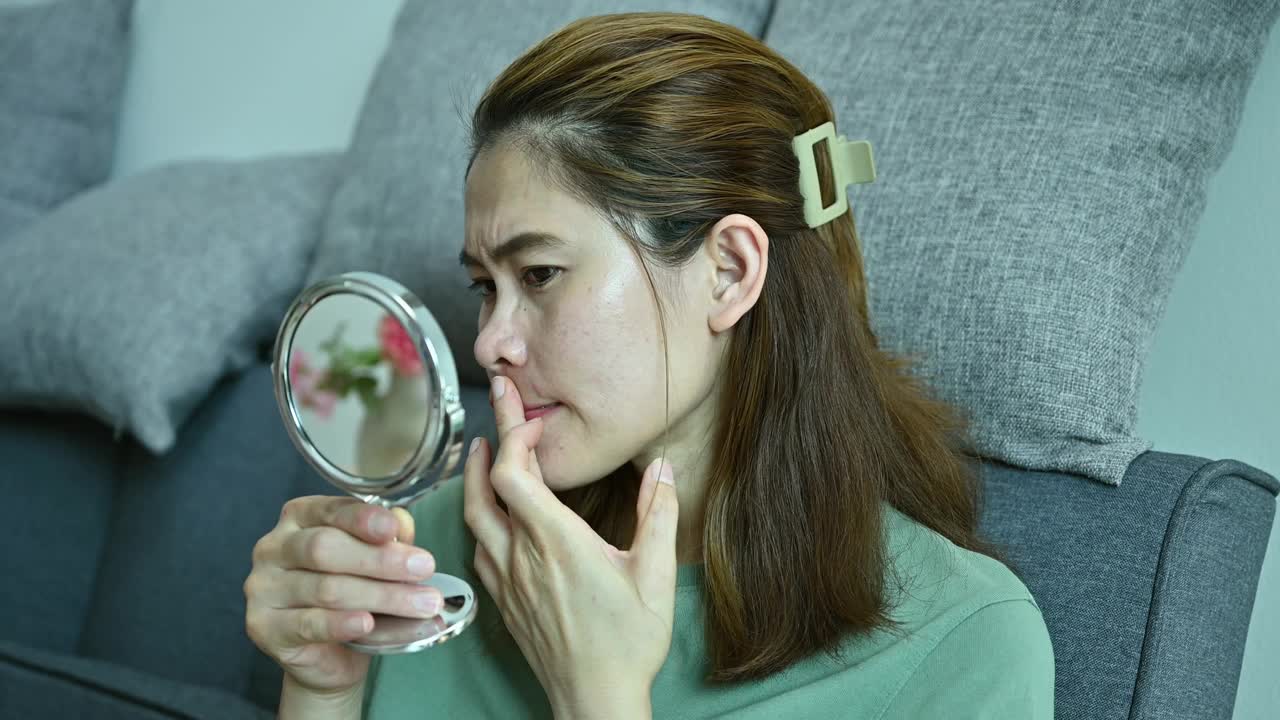 亚洲女性通过迷你镜子看到自己脸上出现痘痘和雀斑时，担心自己的脸。视频下载