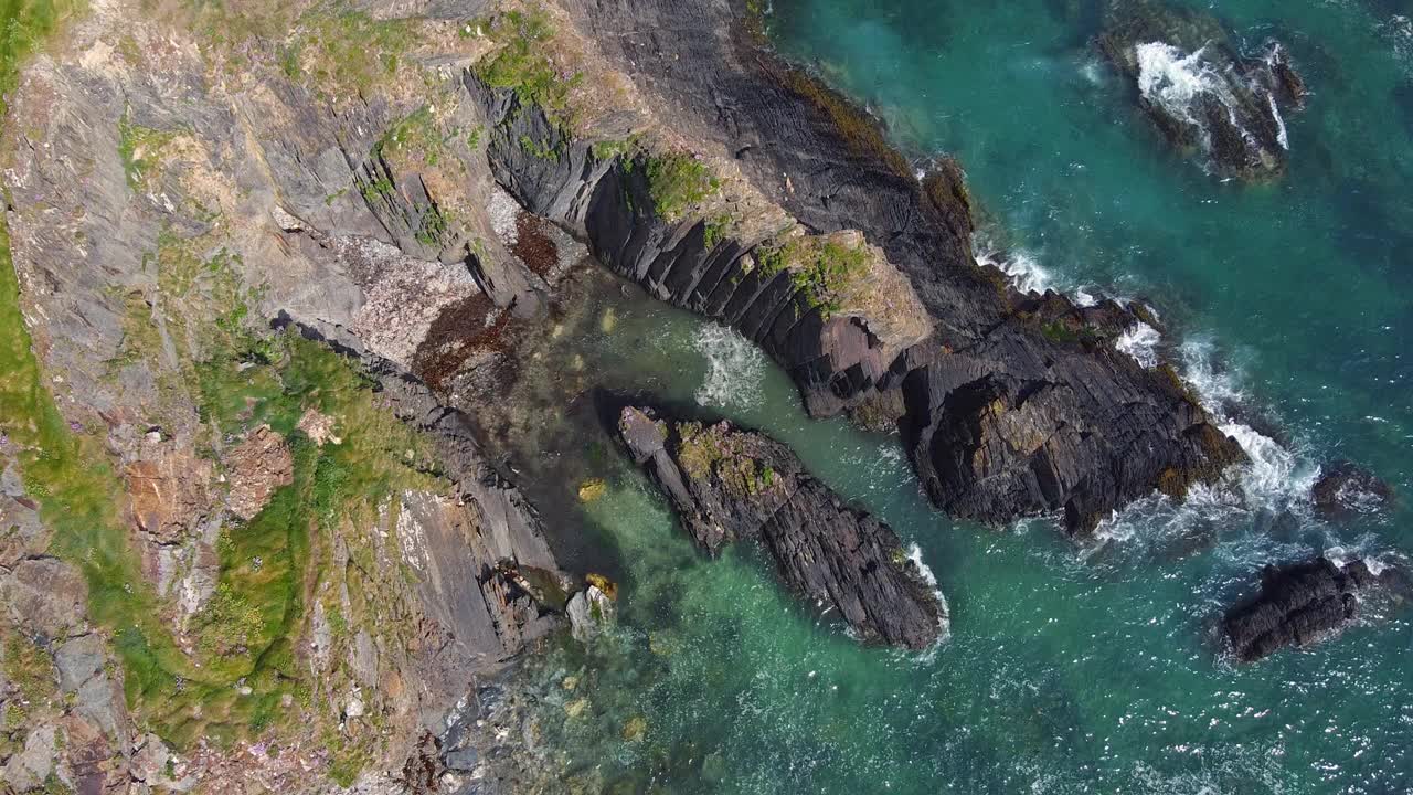 从岩石海岸线上俯瞰的景色。深灰色的岩石与蓝绿色的海水和白色的浪花形成鲜明的对比。岩石经过风化和侵蚀，形成了各种形状和纹理。视频下载