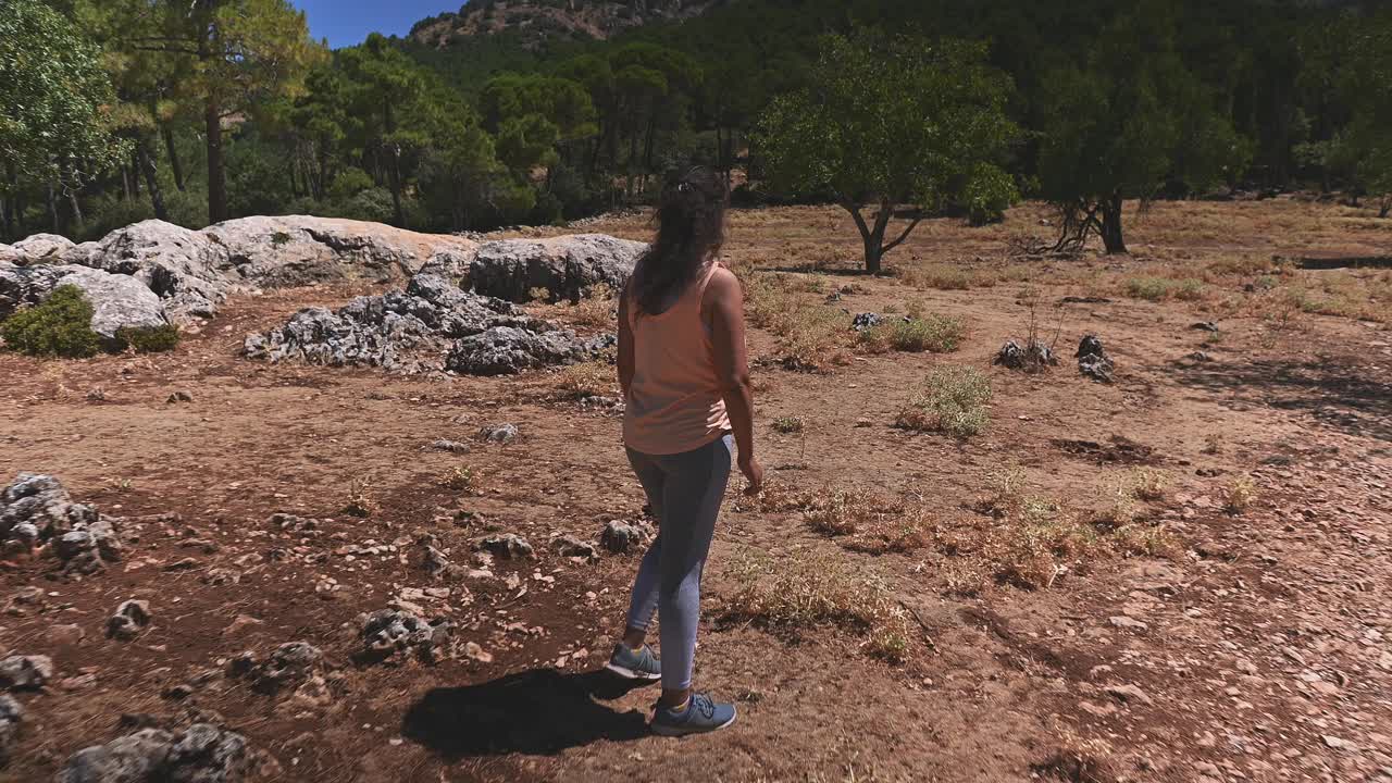 在一个阳光明媚的日子里，一个女人在大自然和树木环绕的风景优美的岩石地带享受宁静的徒步旅行，拥抱户外冒险和宁静的环境视频素材