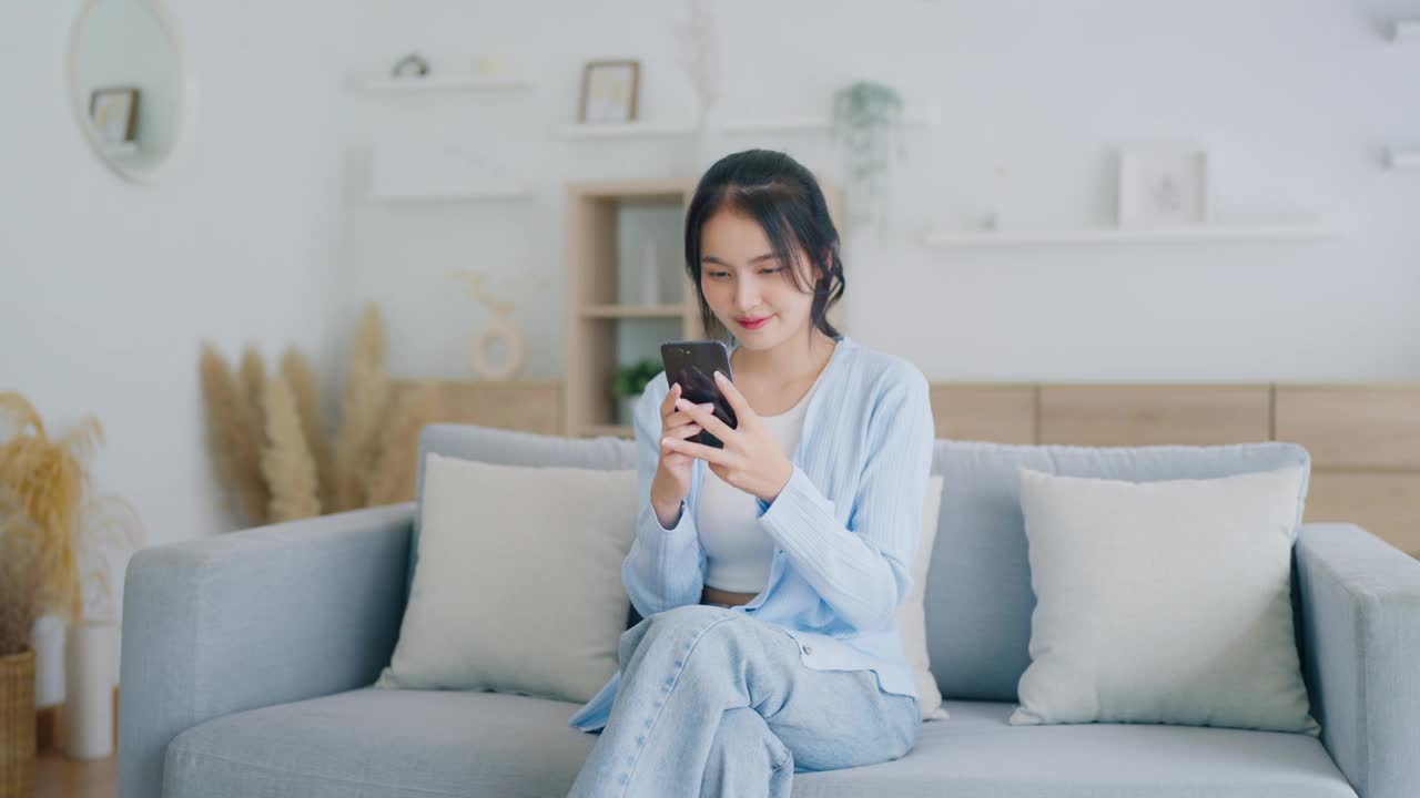 微笑的年轻漂亮的亚洲女人坐在舒适的沙发上，在客厅里使用有趣的手机应用程序。女人在家，网上购物，给朋友发短信，在社交媒体上发帖视频下载