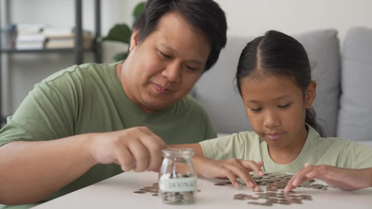 一个亚洲小女孩在家里和她的父亲一起学习投资积蓄，收集罐子里的硬币视频下载