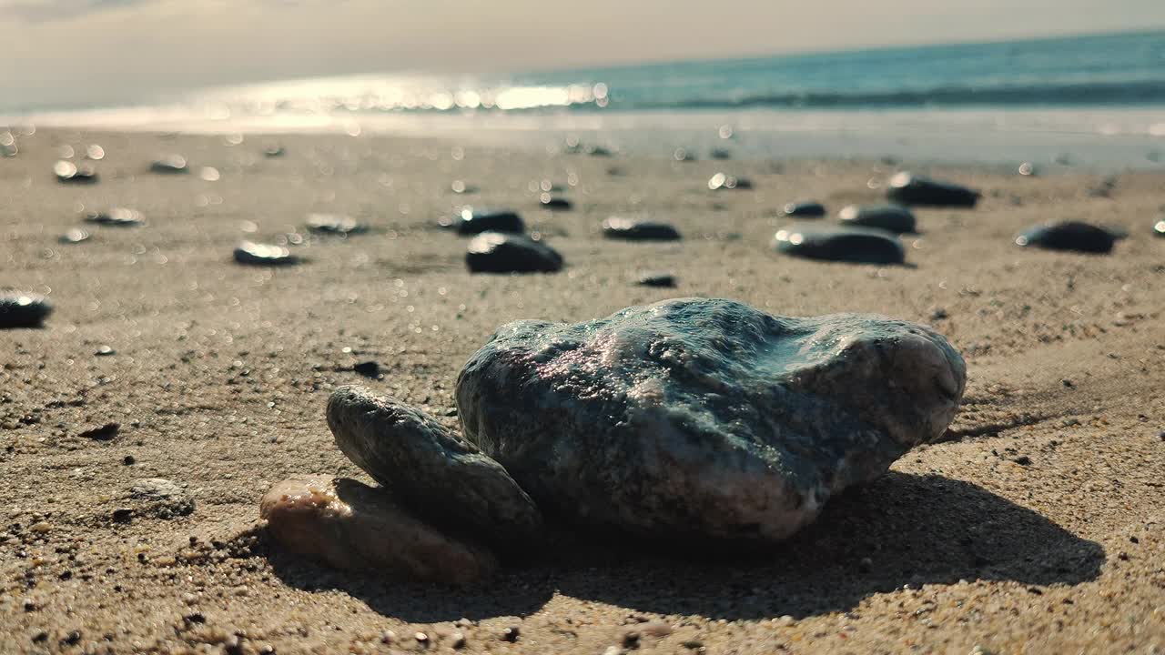 沙滩上闪闪发光的岩石特写，背景是阳光反射在波光粼粼的海洋上，唤起了一个宁静的海边早晨视频下载