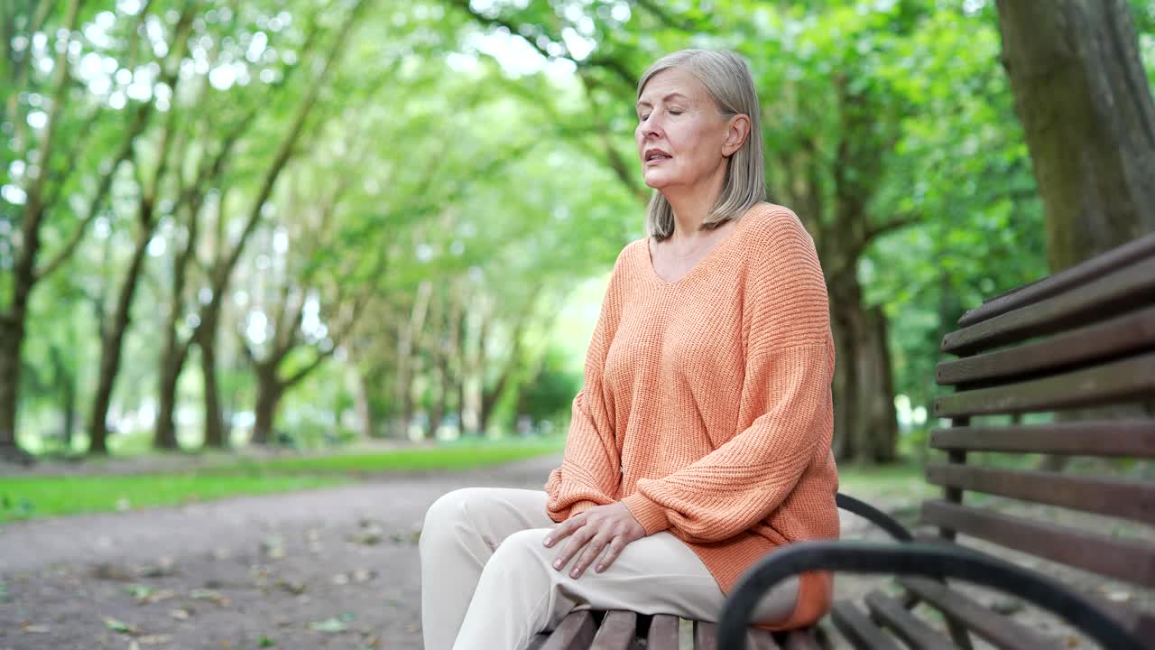 一位白发苍苍的老年女性坐在城市公园街道的长椅上，忍受着严重的膝关节疼痛。视频下载