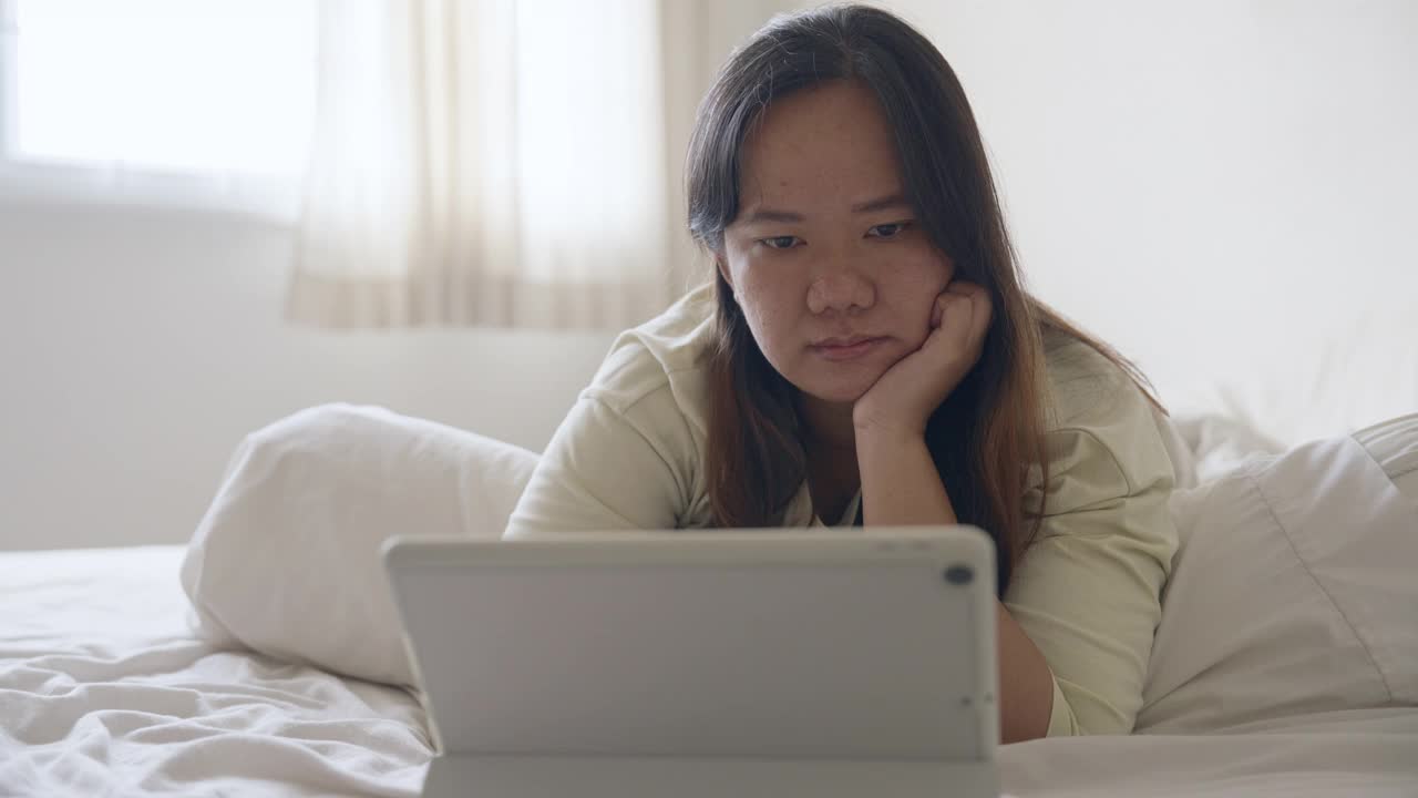 年轻的亚洲女子躺在床上，用数码平板电脑阅读网上的内容视频下载