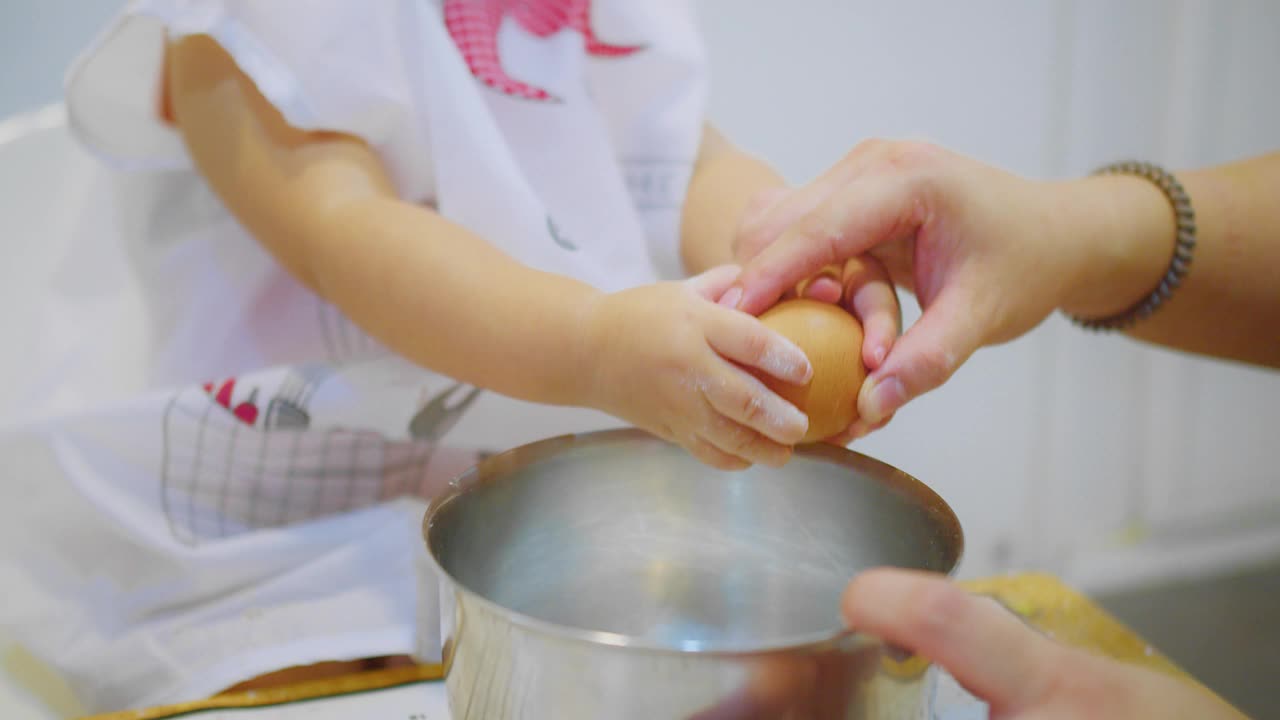 在明亮的厨房里，一个小孩正在和父母一起学习烹饪，他正在把一个鸡蛋打到一个搅拌碗里。视频下载