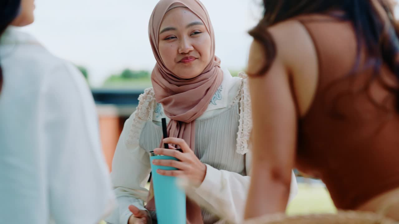 三名年轻女子，其中一名戴着头巾，坐在户外的毯子上，在一个放松的公园环境中享受着交谈和亲密。视频素材