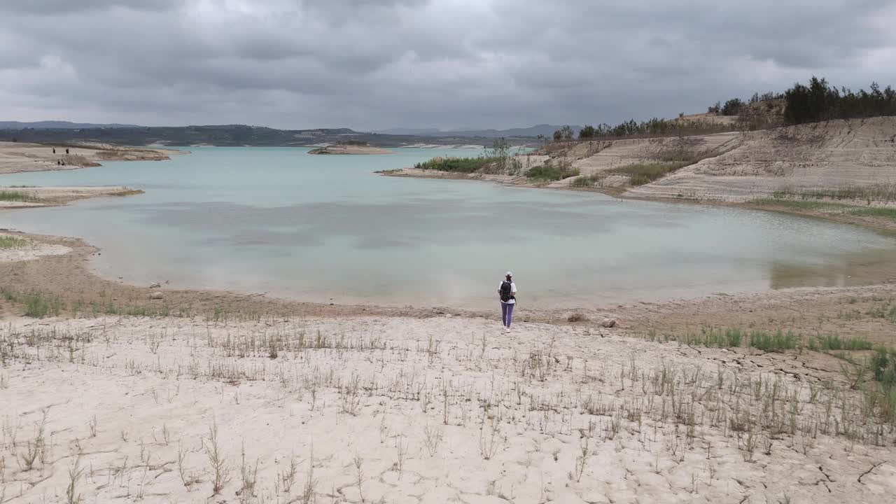 空中飞行。一名女子背着背包走向干涸的湖泊，背景是山丘。鸟瞰美丽的山景视频下载