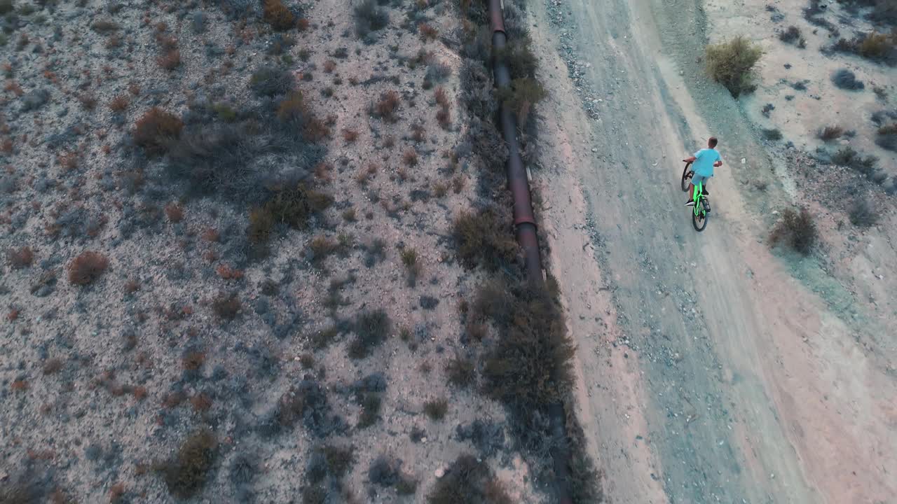 空中飞行。无人机电影镜头骑自行车的人骑山地车沿着尘土飞扬的道路旁边的大水管。鸟瞰美丽的山景视频下载