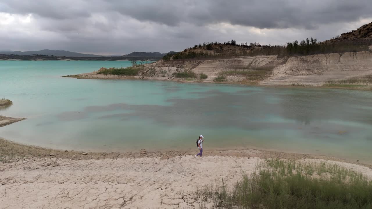 空中飞行。阴天，背着背包的女子走在美丽的绿松石湖畔。鸟瞰美丽的山景视频下载