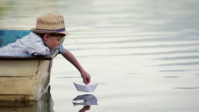 戴着柳条帽的男孩坐在船上，坐着他的纸船去旅行视频素材