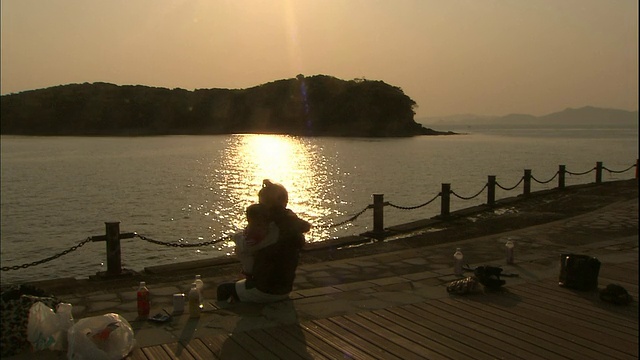 一位母亲抱着她的孩子沿着海岸线走，太阳从水面反射回来。视频素材