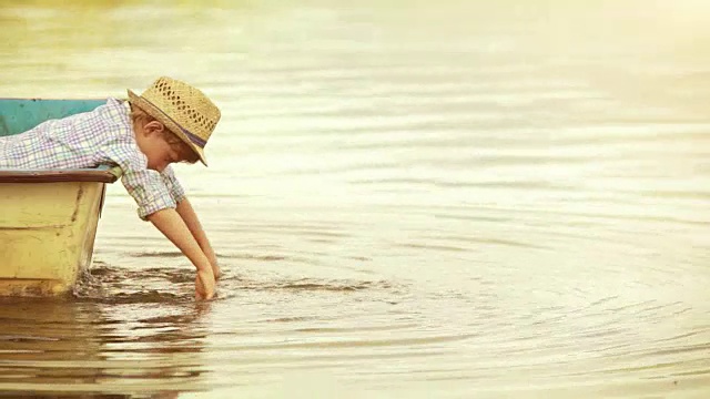 无忧无虑的男孩坐在木船上，双手在水里摇摆视频素材