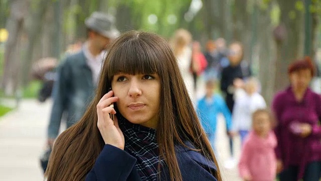 有魅力的年轻女子在蓝色外套在公园的小巷里打电话。在户外用手机聊天的快乐女孩。特写镜头视频素材