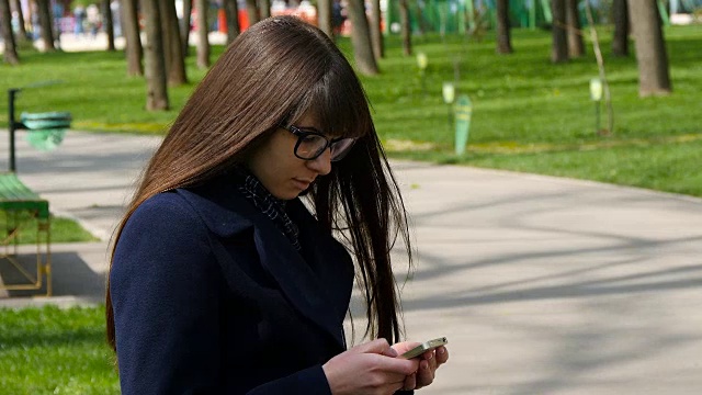 戴眼镜的漂亮女人在公园户外使用手机-细节。年轻迷人的快乐女孩放松在一个城市公园和使用手机。她看起来很高兴，很满足视频素材