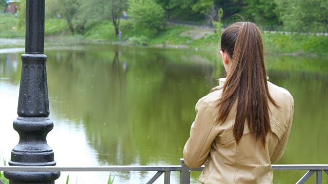 年轻女孩喝着一次性杯子里的外卖咖啡的背影。女人若有所思地望着河，端着一杯茶暖暖身子视频素材