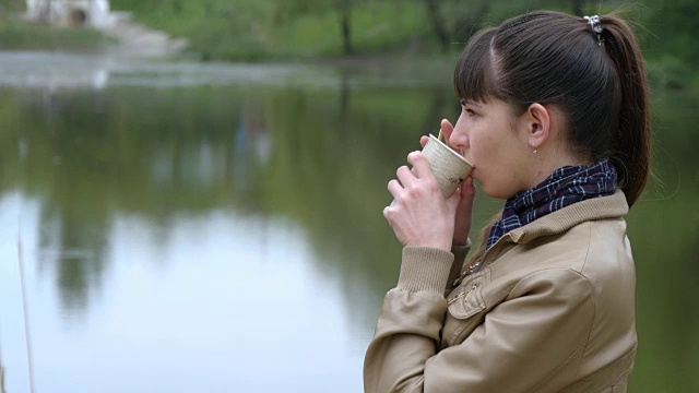 美丽的年轻女孩站在河堤上，喝着一次性杯子里的外卖咖啡视频素材