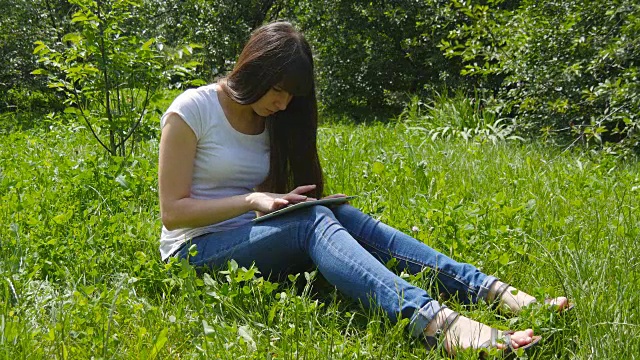 年轻的白人黑发女孩坐在草地上的草地上，在公园里使用数字平板电脑。快乐的女人看着平板电脑屏幕视频素材