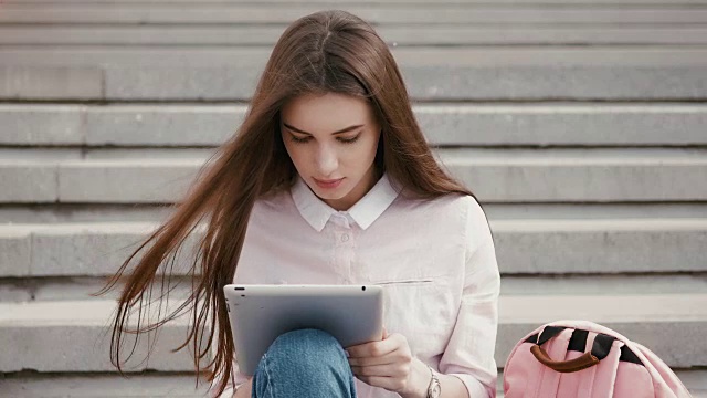 年轻漂亮时髦的女孩坐在台阶上，手握平板电脑。女生在社交网络上与朋友交流视频素材