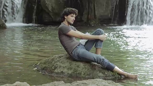 徒步旅行者，探险家在溪流中间的一块石头上休息，瀑布在视频素材