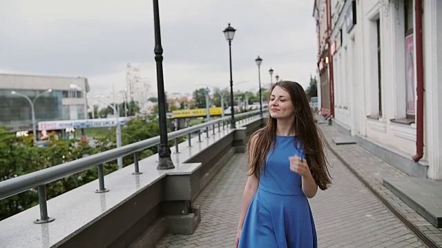 美丽的年轻女子穿着蓝色的裙子拿着购物袋走在城市里，环顾四周，慢悠悠地拍了拍视频素材