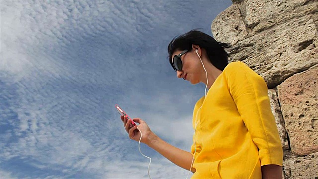 美女用智能手机耳机听音乐，背景是天空和古城废墟视频下载