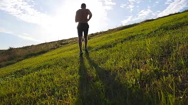 年轻人跑过绿色的山在蓝天的背景。日落时分，男运动员在大自然中慢跑。运动跑步者在日出时在户外上坡慢跑。越野训练。慢动作视频素材