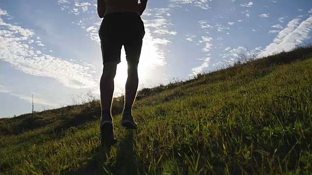 年轻人跑过绿色的山在蓝天的背景。日落时分，男运动员在大自然中慢跑。运动跑步者在日出时在户外上坡慢跑。越野训练。慢动作视频素材