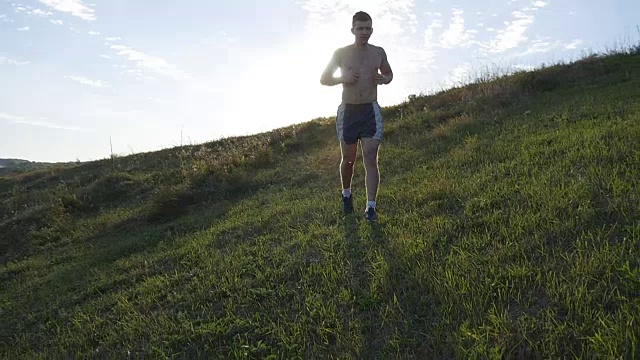 年轻人跑过绿色的山在蓝天的背景。日落时分，男运动员在大自然中慢跑。运动跑步者在日出时在户外上坡慢跑。越野训练。慢动作视频素材