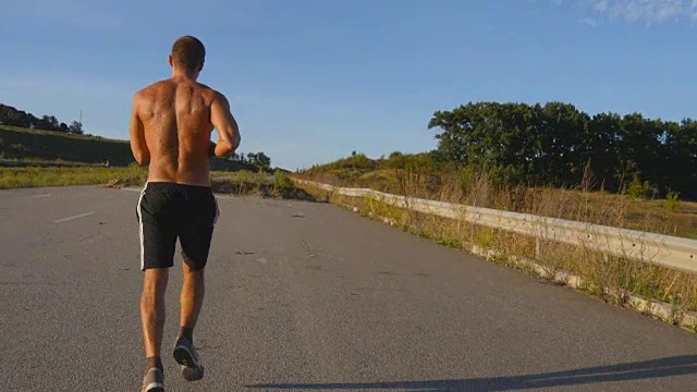 短跑运动员在高速公路上慢跑。男子运动运动员夏季户外训练。年轻强壮的肌肉男锻炼在农村道路上锻炼期间。积极健康的生活方式。近距离视频素材